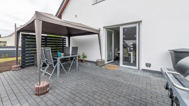 Eine Terrasse mit überdachter Sitzgelegenheit, einem Grill und einer Glastür zu einem Innenbereich.