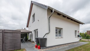 Ein zweistöckiges Haus mit weißer Fassade, einem grauen Dach und einem grauen Regenrinnensystem. Auf der linken Seite steht eine kleine Hütte und eine gepflasterte Auffahrt führt zum Haus.