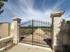 Une entrée fermée par un portail en fer forgé, flanquée de piliers en pierre, qui mène à un chemin dallé.