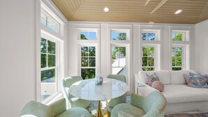 Une pièce avec une table ronde en verre, des fauteuils vert clair, un canapé blanc agrémenté de coussins colorés et de grandes fenêtres donnant sur un paysage verdoyant.