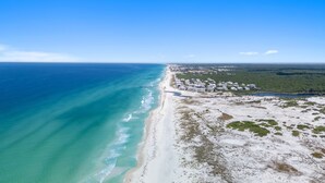Vue aérienne d’une zone côtière comprenant une plage, un petit village avec des maisons et une forêt en arrière-plan.