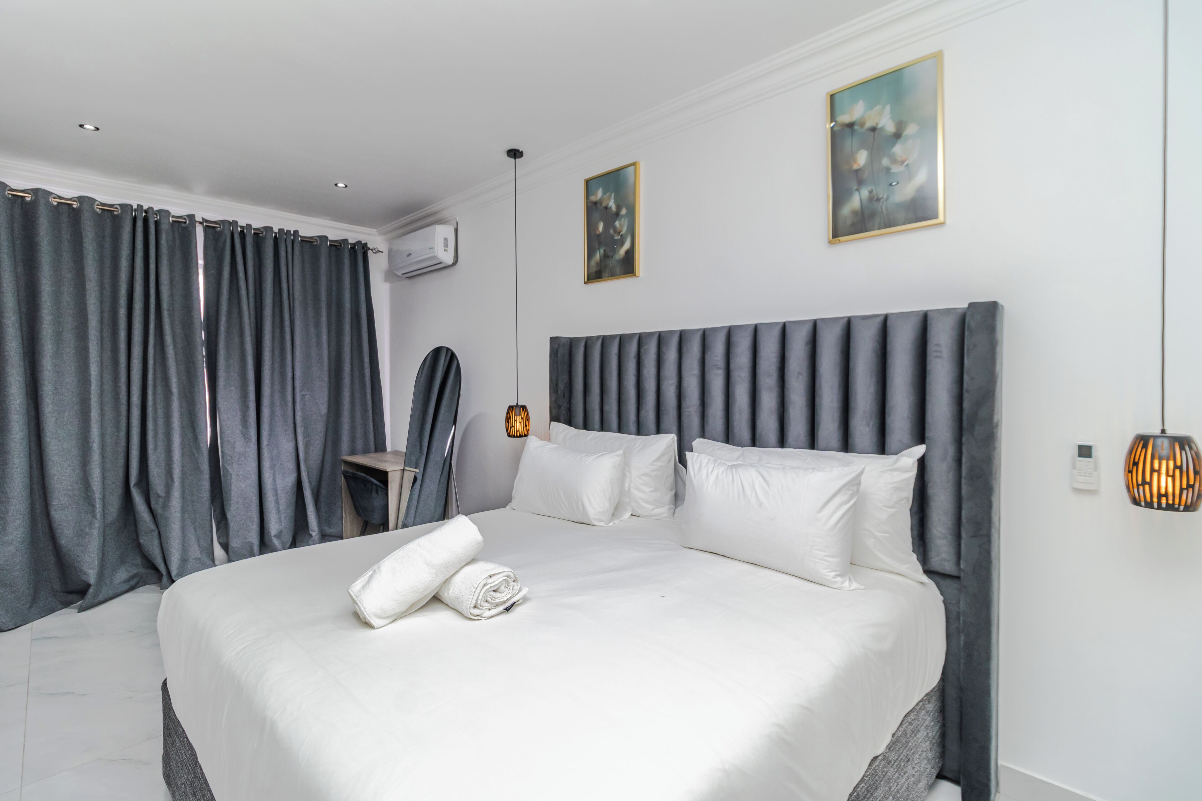 A modern hotel room with a large bed, grey curtains, a bedside table, and a wall-mounted air conditioner.