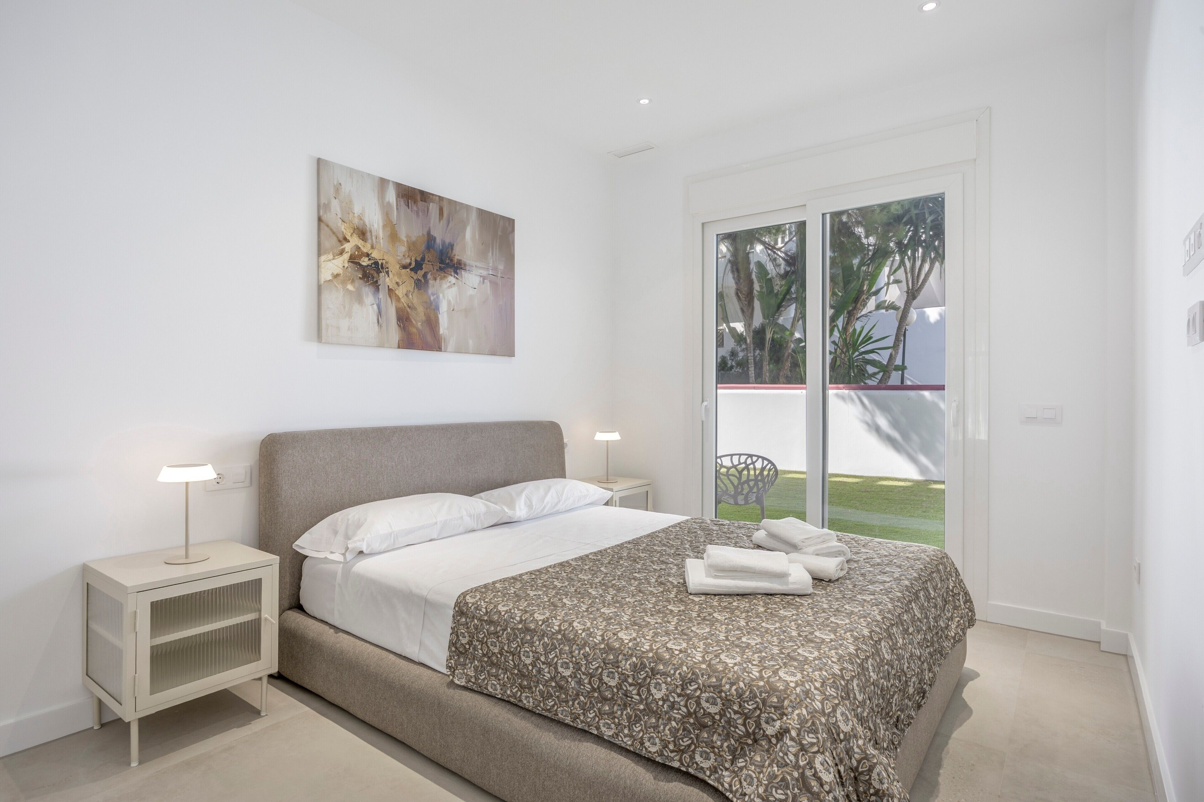 Une chambre moderne avec un lit, des tables de chevet, une lampe et une grande fenêtre donnant sur un paysage verdoyant.