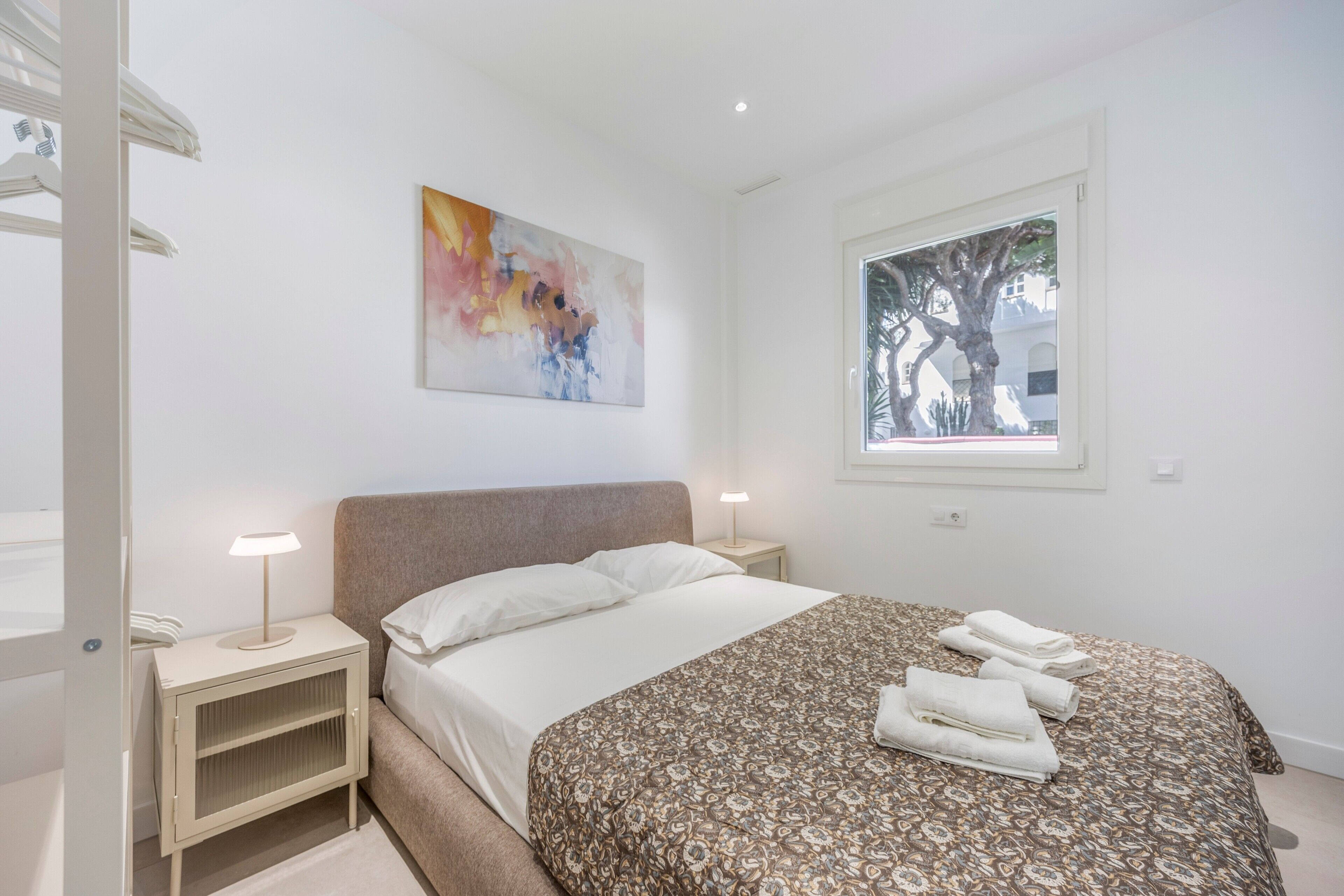 Une chambre à coucher avec un lit, une table de chevet, une lampe, une fenêtre donnant sur un arbre et un tableau au mur.