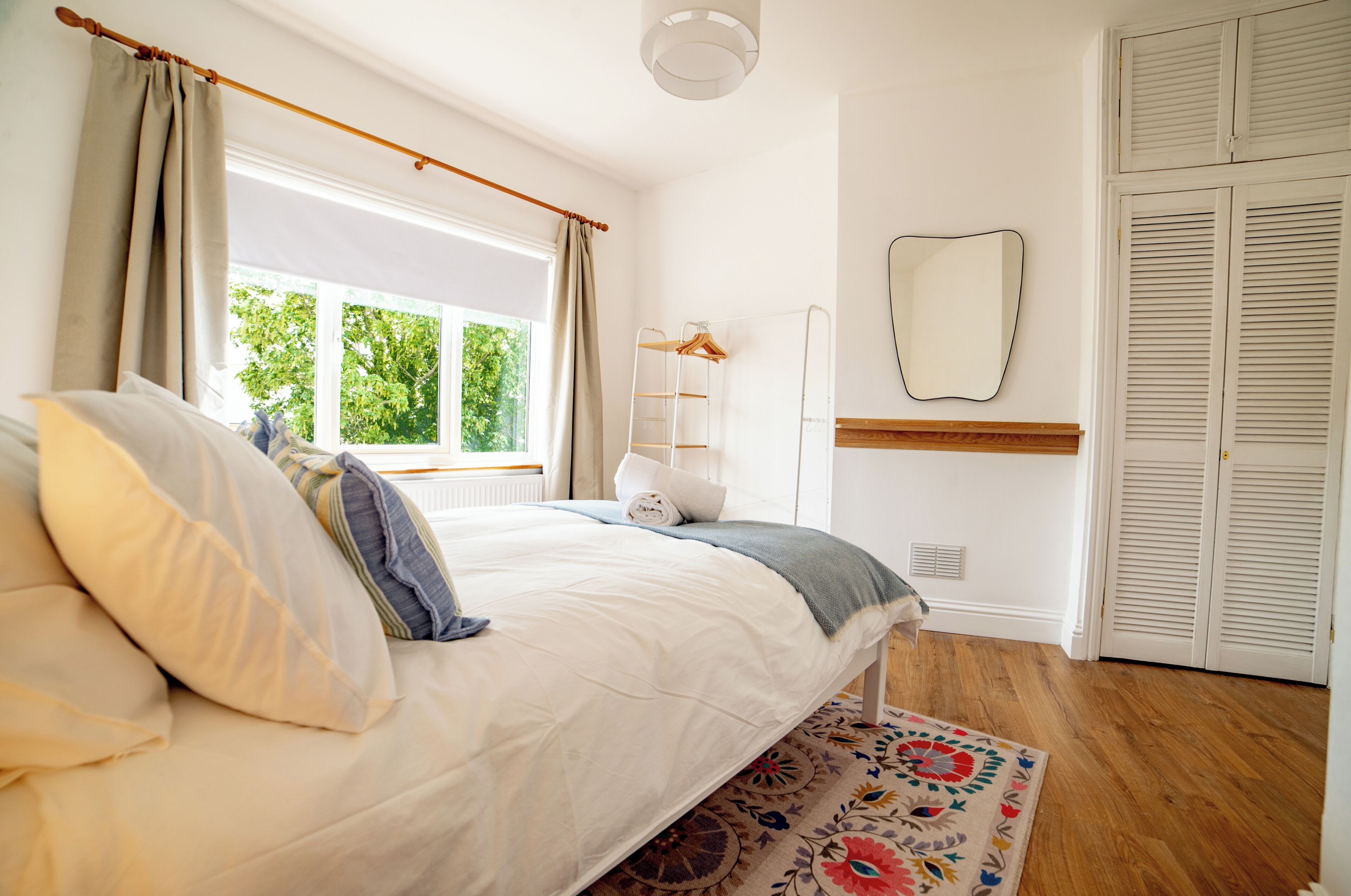 Ein Schlafzimmer mit Bett, Fenster mit Vorhängen, Holzboden, Spiegel und Kleiderschrank.