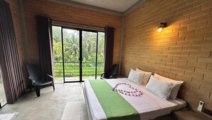 Quarto de hotel com uma cama, duas cadeiras, vista por uma porta de vidro e parede de tijolos.