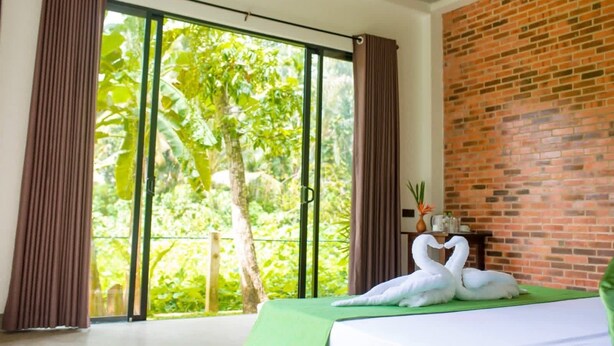 Quarto com uma cama, um cobreleira verde, parede de tijolos e vista para o verde através de grandes janelas.
