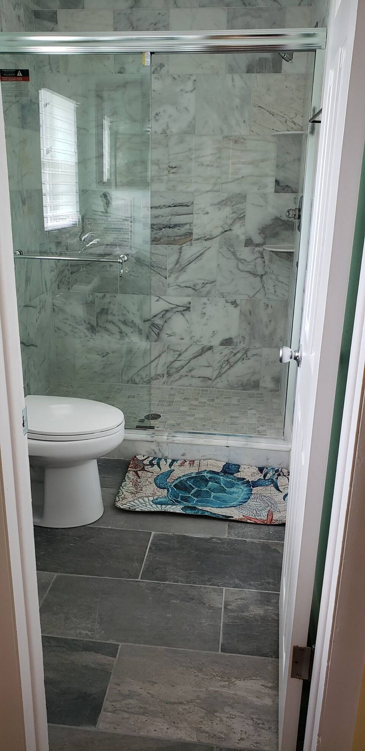 A bathroom with a glass shower enclosure, a toilet, and a patterned mat.
