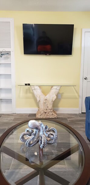 A living room with a flat-screen TV mounted on a yellow wall, a glass coffee table with an octopus sculpture, and a decorative shell piece. - Modern Beach Retreat - Mermaid Cottage (St. Augustine)