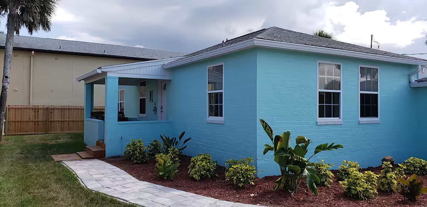 A single-story house with a blue exterior, a front porch, and a small garden with various plants.