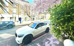 Ein weißes Auto steht auf einer Straße, im Hintergrund sind Gebäude und ein Baum zu sehen.