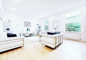A modern living room with a white sofa, a white coffee table, and a large window with a view of greenery. - Designer Luxe Apartment in Brackenbury Village, A/C, 2 large beds (London)
