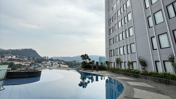 A high-rise building with a swimming pool, a view of the cityscape, and a hilly background. - 2-bedroom apartment in Bandar Lampung with Mall Access (Bandar Lampung)