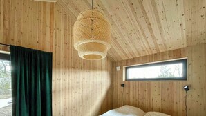Une pièce aux murs en bois, un lit avec une literie blanche, un luminaire suspendu en fibres tressées, une fenêtre et un rideau.