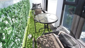 En balkon med et glasbord og fletstole, et område med grøn kunstgræs og udsigt over bybilledet.