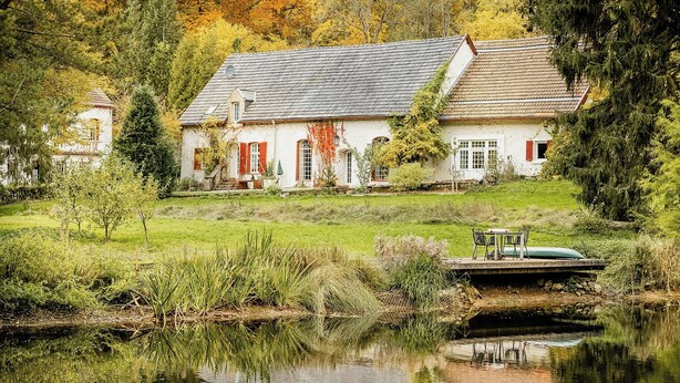 Ein weißes Haus mit einer Steinsbrücke über einem Teich, umgeben von Grünflächen und Bäumen.