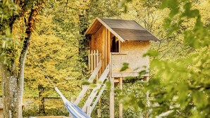 Ein hölzernes Baumhaus mit einer Leiter, umgeben von Bäumen und einer Hängematte.