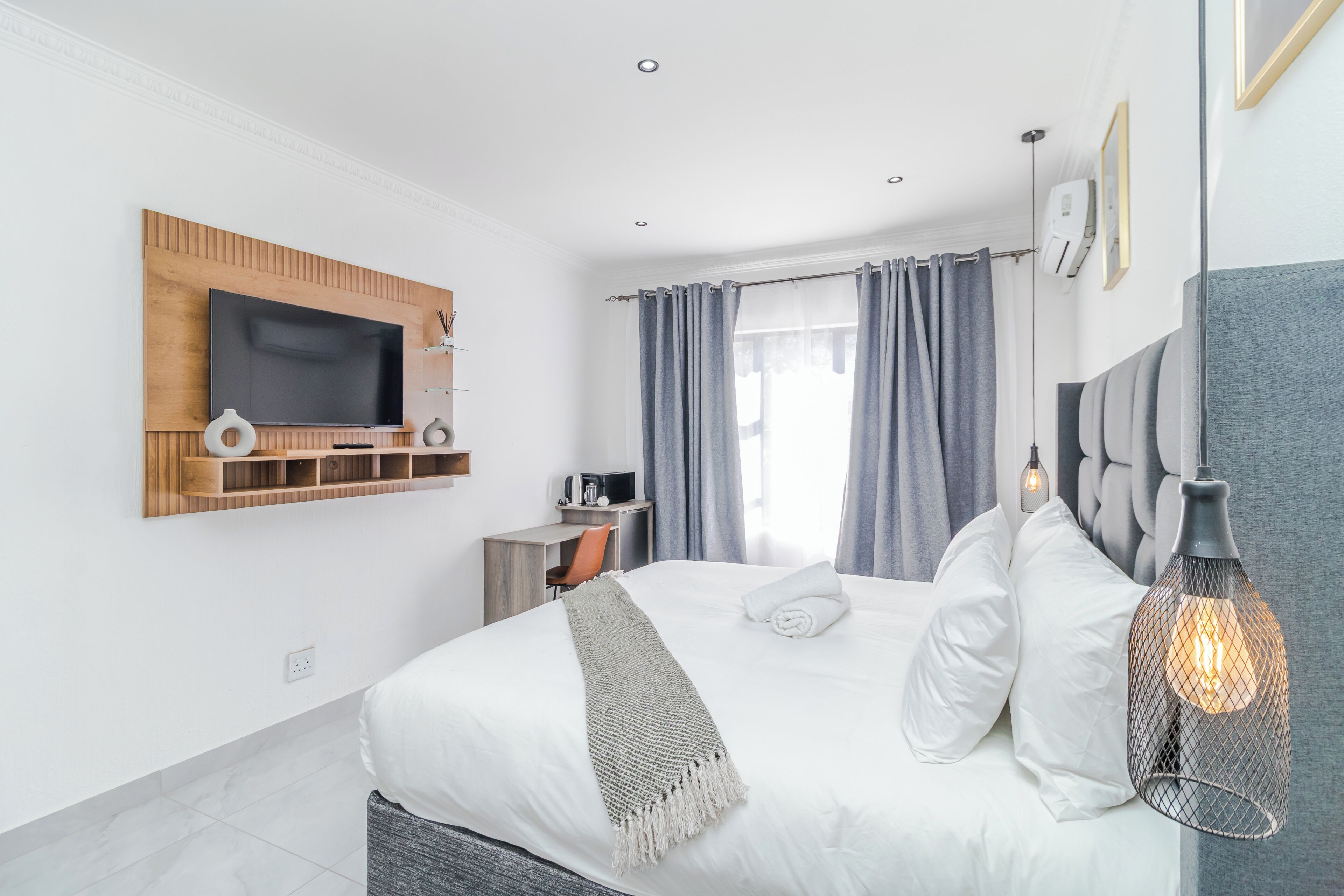 A modern hotel room with a large bed, a TV mounted on the wall, a desk with a chair, and a hanging light fixture.