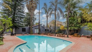 A swimming pool with a tiled edge, surrounded by palm trees and a patio area with tables and chairs. - Boutique Hotel-Style Room In Ferndale-D4 (Randburg)