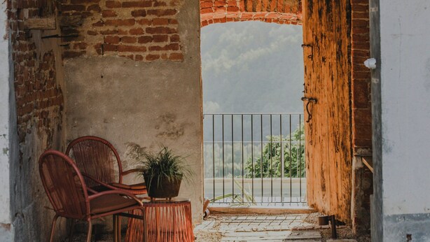 An arched brick doorway leading to a balcony with a view, a wicker chair, and a potted plant. - Noble residence with pool in the Langhe (Cherasco)