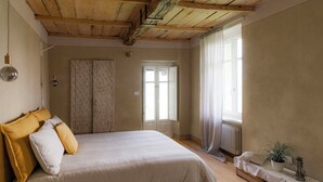 A bedroom with a bed, a wooden ceiling, a radiator, a window with curtains, and a bedside table with a plant. - Noble residence with pool in the Langhe (Cherasco)