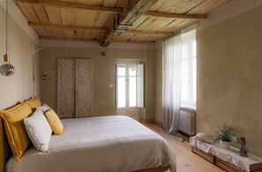 Ein Schlafzimmer mit einem Bett, Holzboden, Heizkörper, einem Fenster mit Vorhängen und einem Nachttisch mit Pflanze.