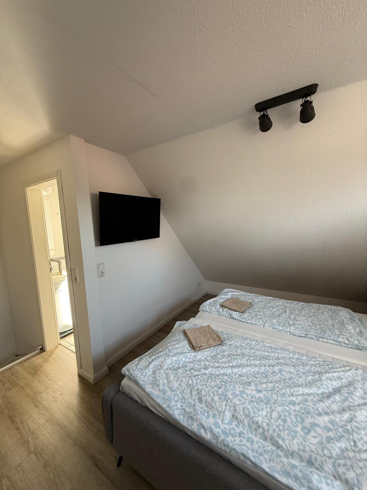A room with a bed, a flat-screen TV mounted on the wall, and a ceiling light fixture.