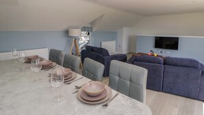 A modern living room with a dining area, grey sofas, a TV mounted on the wall, and a ceiling light fixture. - Rosemary Cottage 2 (Tywyn)