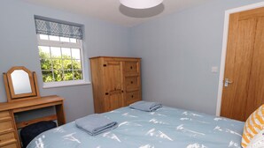 A bedroom with a bed, a wooden dresser, a desk with a mirror, and a window with a view of greenery. - Rosemary Cottage 2 (Tywyn)