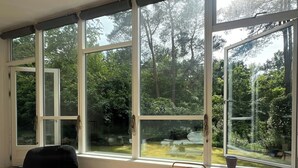 A room with large windows, a black chair, a table with a vase, and a red bag.