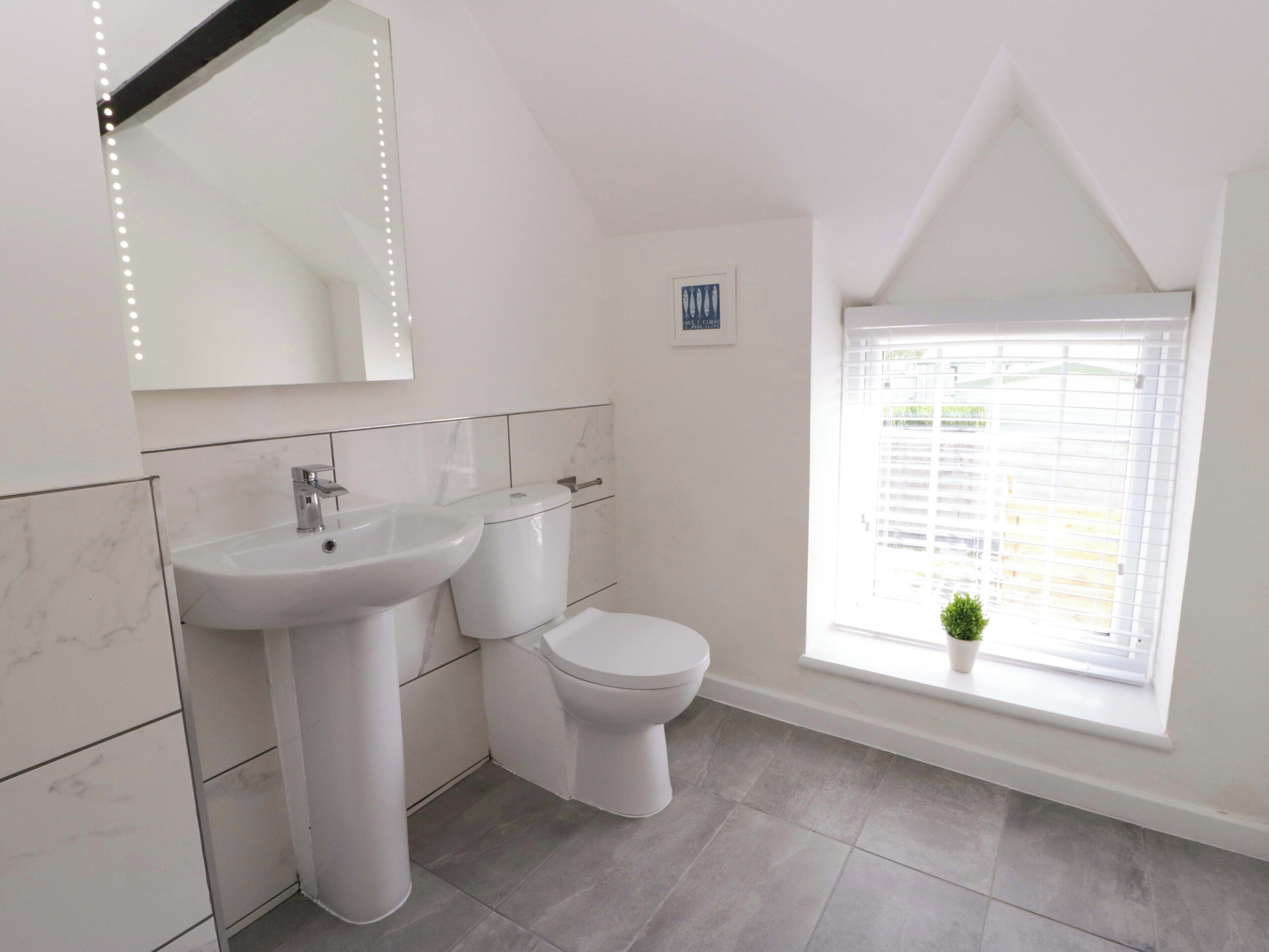 Een badkamer met een wit wastafelmeubel, toilet en spiegel. Er is een raam met zonwering en een klein plantje op de vensterbank.