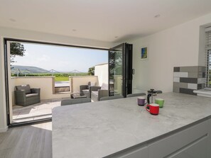 A modern kitchen with a large island, a sliding glass door leading to a patio with outdoor seating, and a view of the countryside. - Rosemary Cottage 1 (Tywyn)