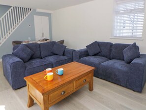 A living room with two grey sofas, a wooden coffee table, and a dining area with a table set for four. - Rosemary Cottage 1 (Tywyn)