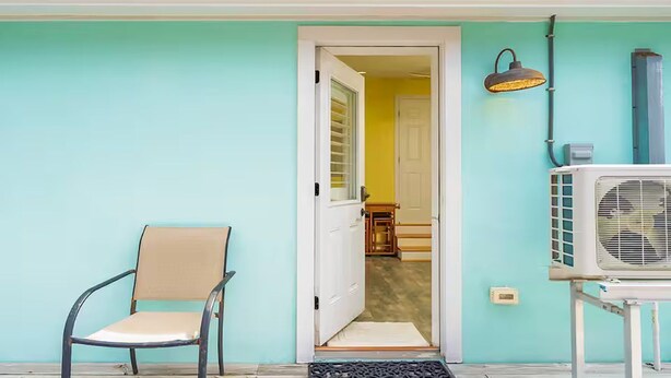 Een wand in blauwgroen, een witte deur die naar de binnenkant leidt, een stoel en een airconditioning.