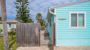 Een turquoise huis met een houten terras, een omheinde tuin en een heldere lucht.
