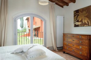 A bedroom with a bed, a dresser, a painting, and a view of an outdoor area with a building and greenery. - Converted barn with sunset views in Tuscany (Reggello)