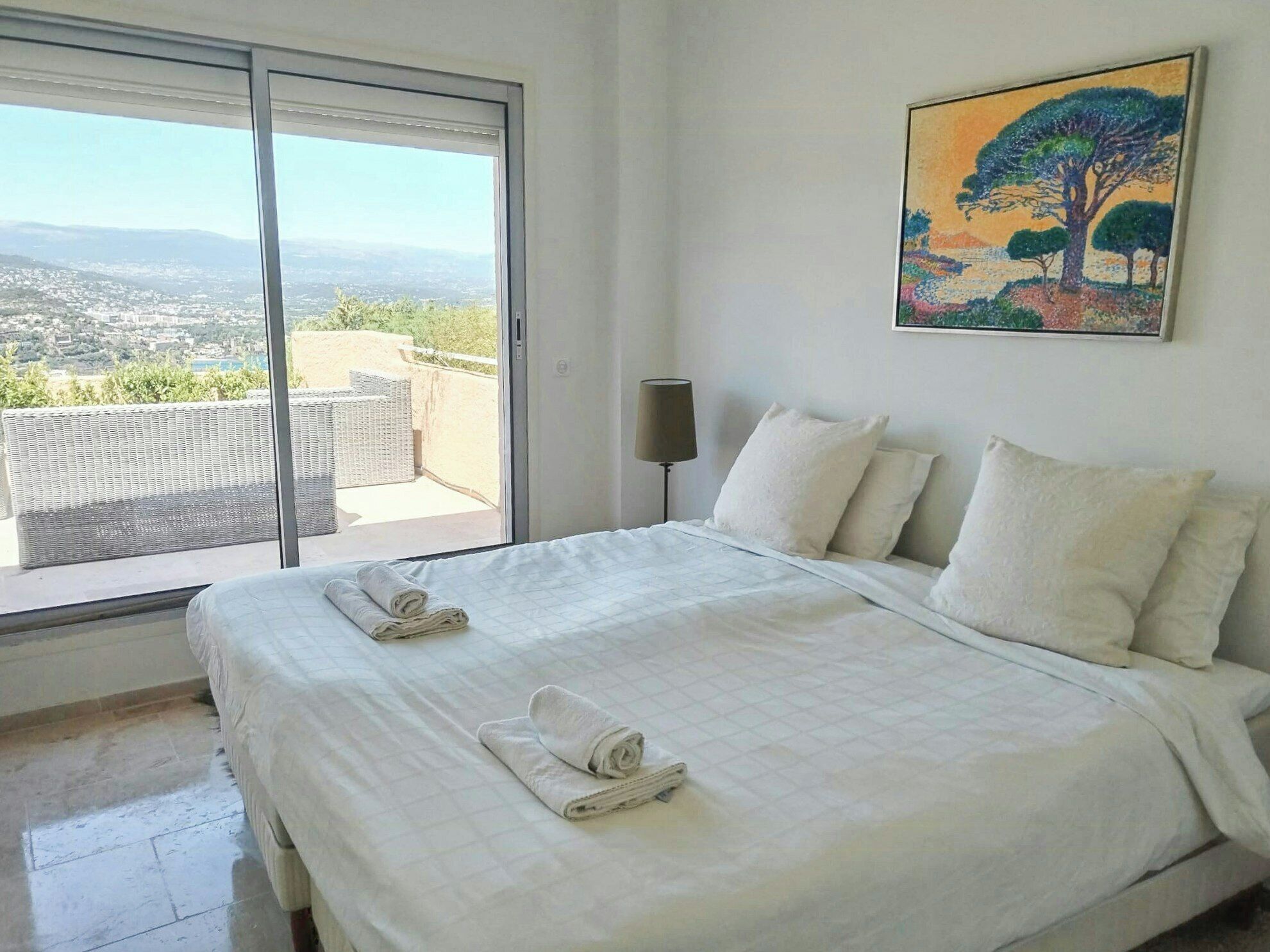 A hotel room with a large bed, a view of the city, and a painting on the wall.