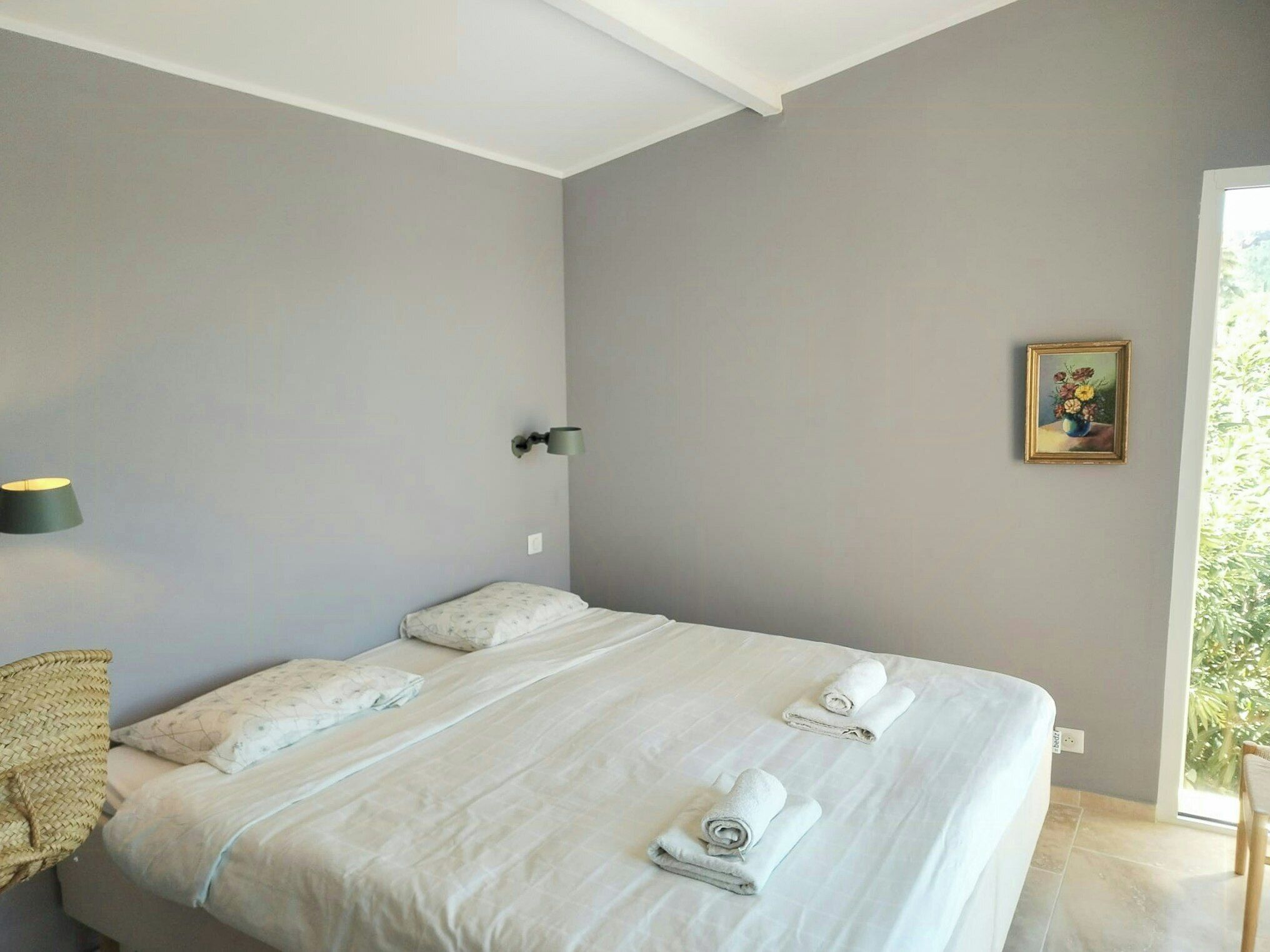 Een slaapkamer met een bed, kussens, een wandlamp, een lijst met een afbeelding en een raam met uitzicht.