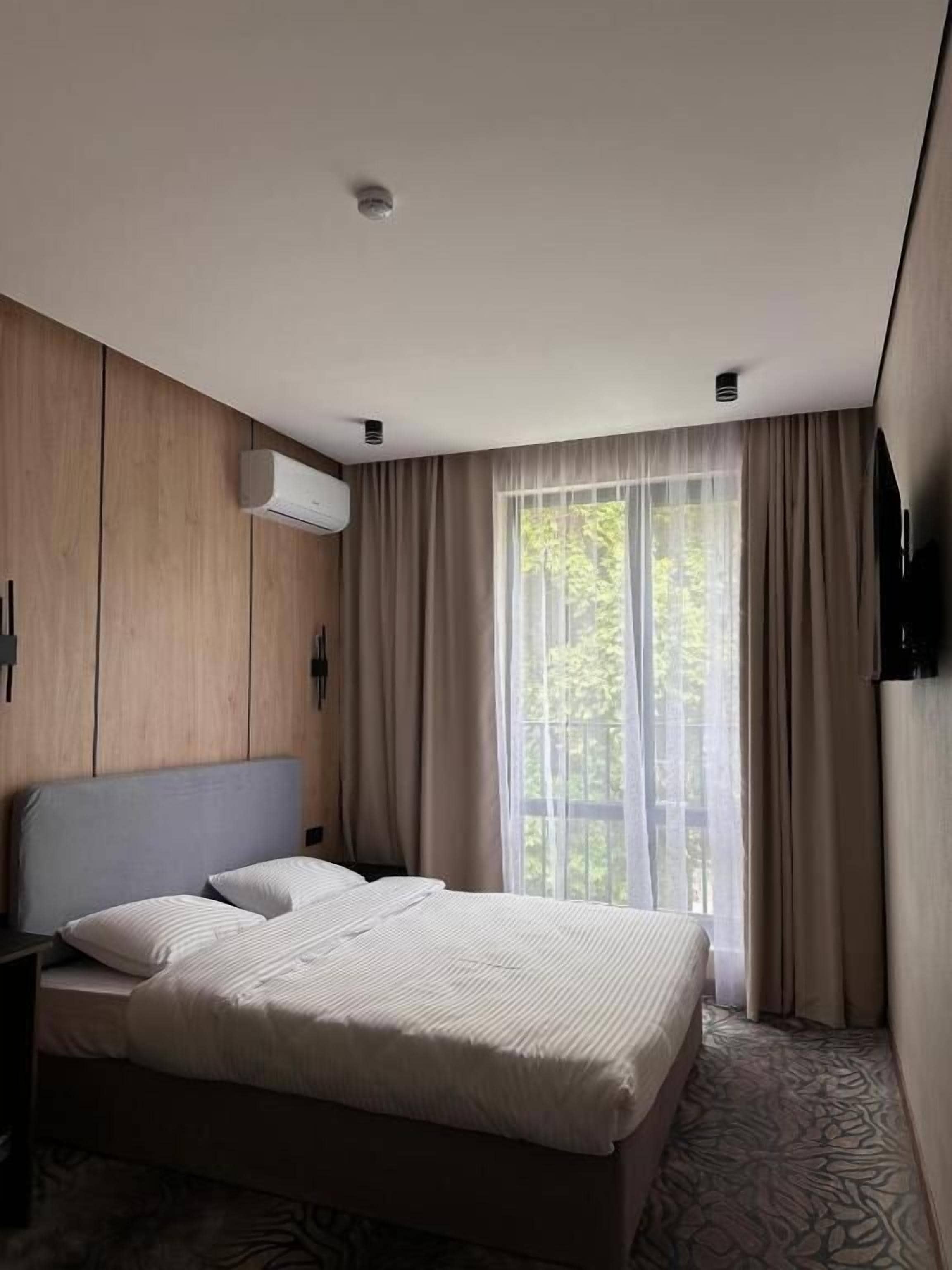 A hotel room with a bed, a TV, an air conditioning unit, and a window with curtains.