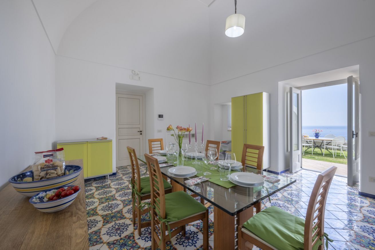 Un comedor con mesa de vidrio, sillas con cojines verdes y vista al mar a través de una puerta abierta.