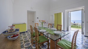 Een eetgedeelte met een glazen tafel, stoelen met groene kussens en uitzicht op zee door een open deur.