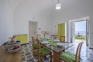 Ein Essbereich mit einem Glastisch, Stühlen mit grünen Kissen und Blick auf das Meer durch eine offene Tür.