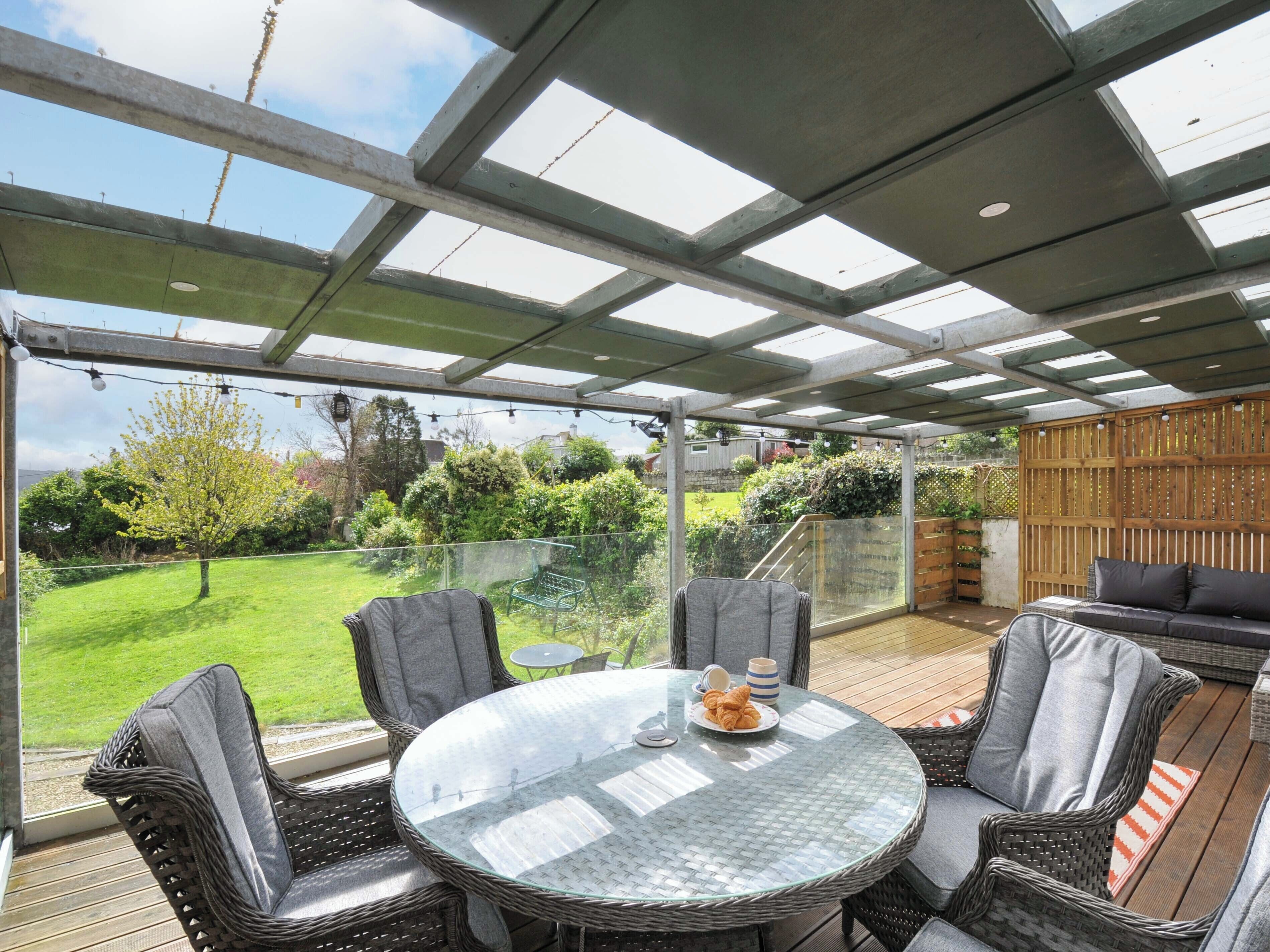 Eine überdachte Terrasse mit Korbmöbeln, einem Glastisch und Blick auf einen grünen Garten.