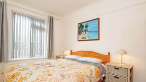Un dormitorio con cama, un cuadro de una playa, una ventana con cortinas y lámparas de noche.