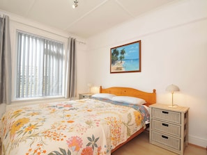 A bedroom with a bed, a painting of a beach scene, a window with curtains, and bedside lamps. - Porthmeor (Wadebridge)