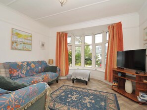 A living room with a sofa, a television on a wooden stand, a small rug, and a window with curtains. - Porthmeor (Wadebridge)