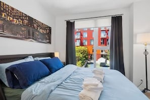 A hotel room with a bed, a painting on the wall, and a view of buildings outside.