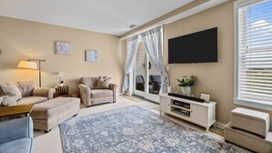 Een woonkamer met een bank, voetenbankje, televisie en een groot raam met jaloezieën.