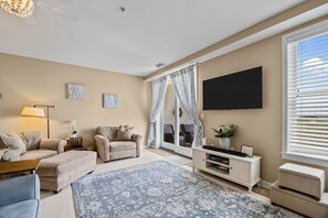 Ein Wohnzimmer mit einer Couch, einem Hocker, einem Fernseher und einem großen Fenster mit Jalousien.