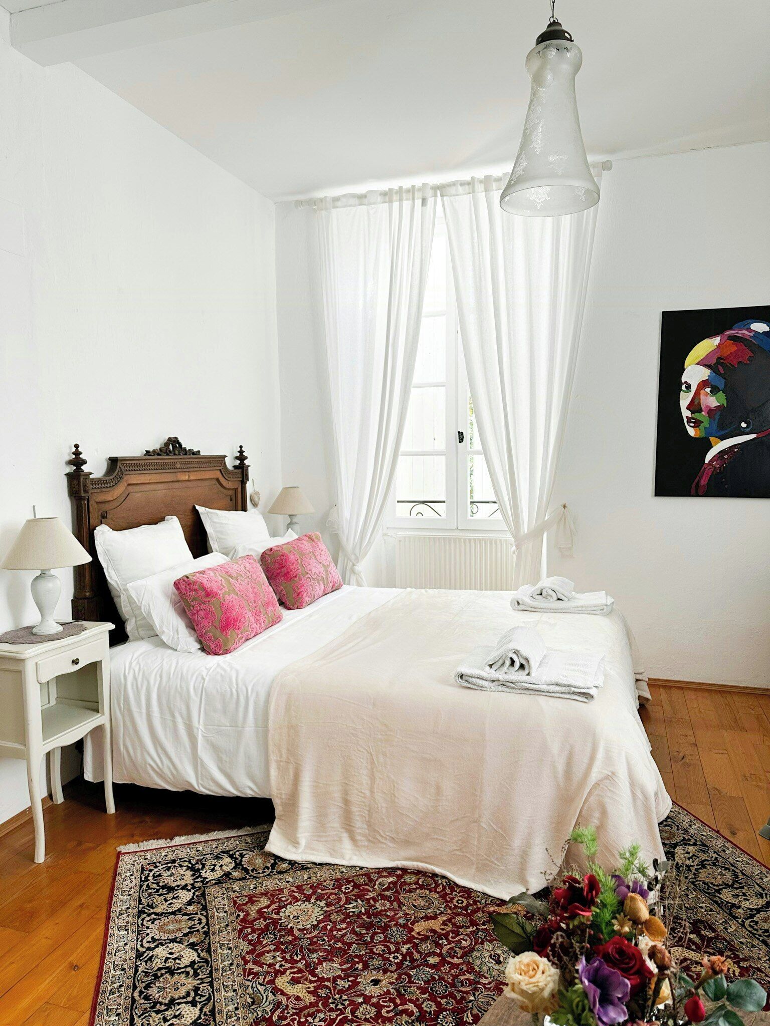 A bedroom with a wooden headboard, white bedding, a bedside table with a lamp, a large window with sheer curtains, a hanging light fixture, and a floral arrangement on the floor.